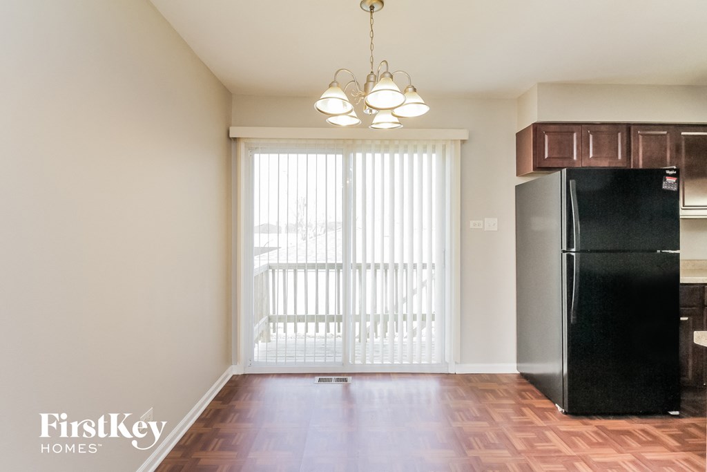 A black fridge sits in a kitchen with a wooden floor and a window with blinds.