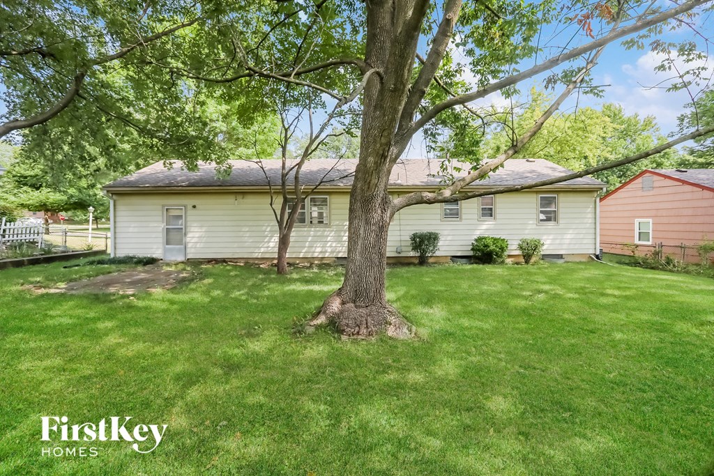 A tree stands in the front yard of a house with a white siding and a red house in the background.
