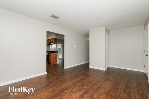 A room with wooden floors and white walls, with a kitchen area visible through a doorway.
