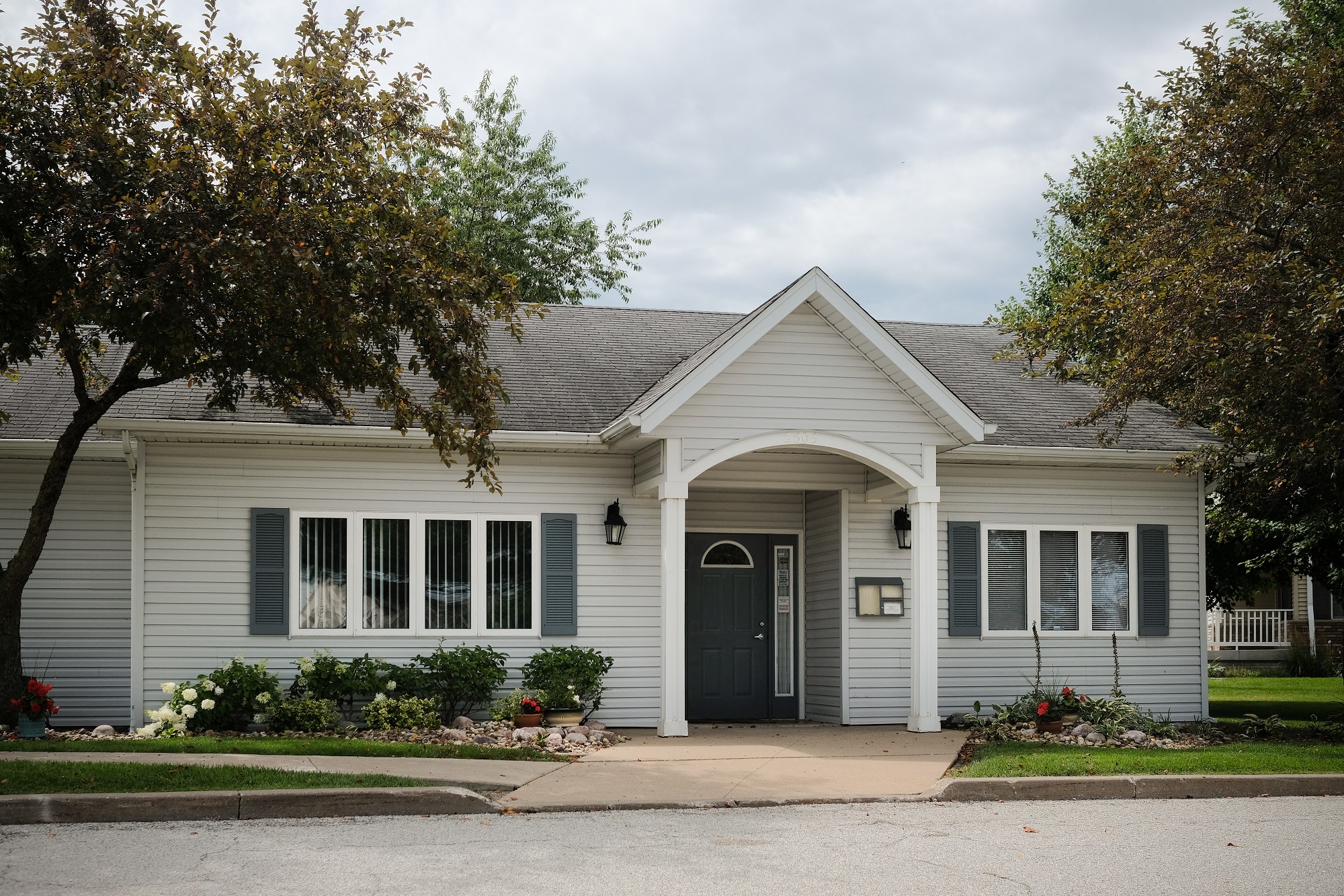 grey siding with mature trees, landscape, bright sky, and leasing office at regency apartments in Bettendorf Iowa