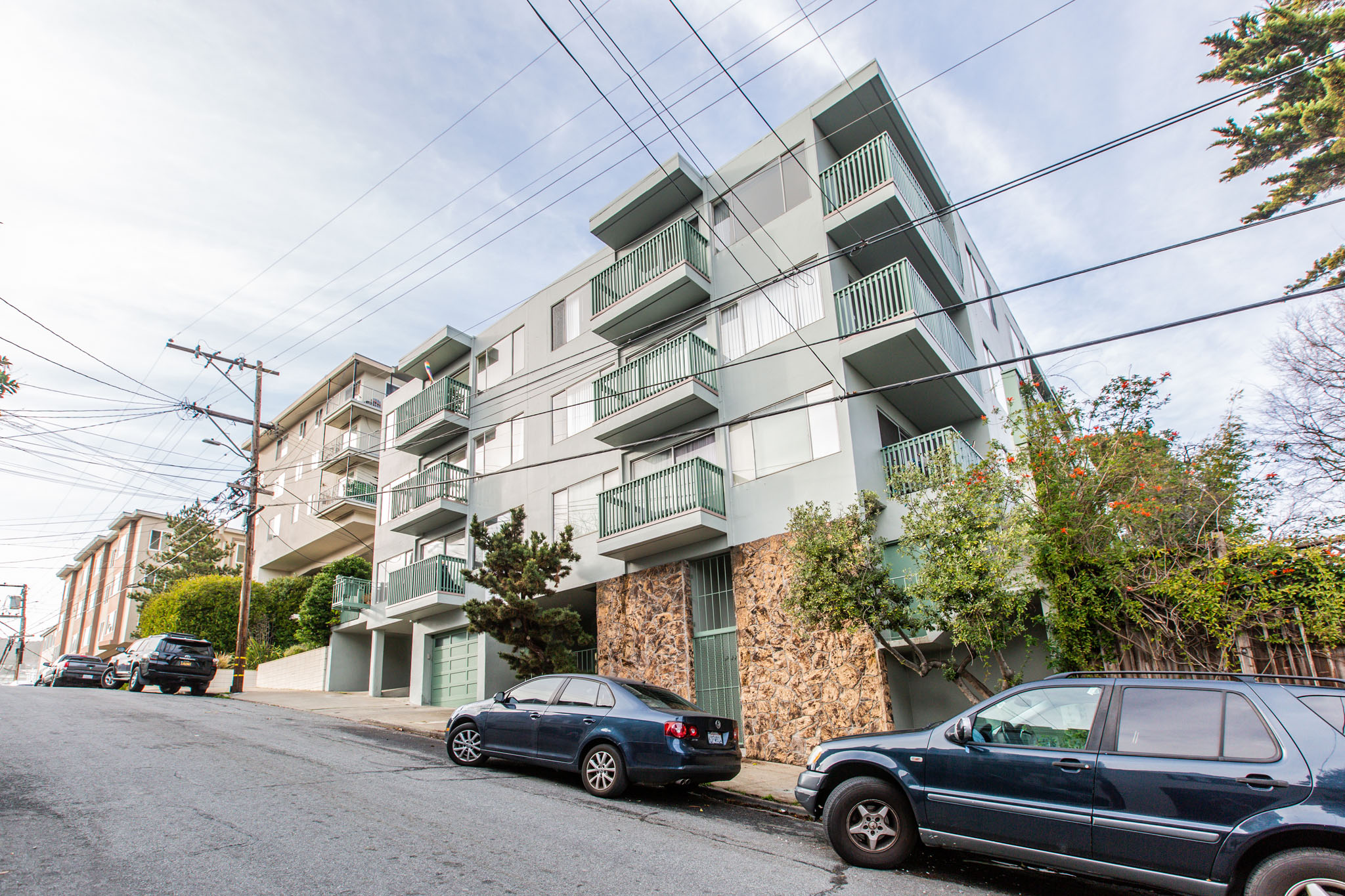 A street view of a residential area with cars parked on the side of the road and apartment buildings in the background.
