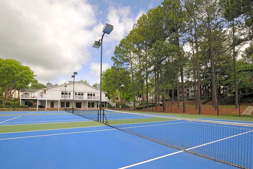 A tennis court with a blue surface and white lines is surrounded by trees and a building.