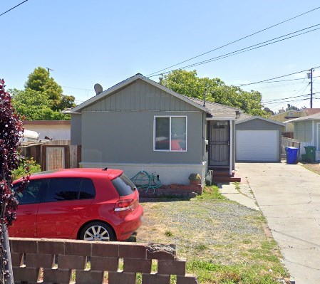 A red car is parked in front of a grey house.