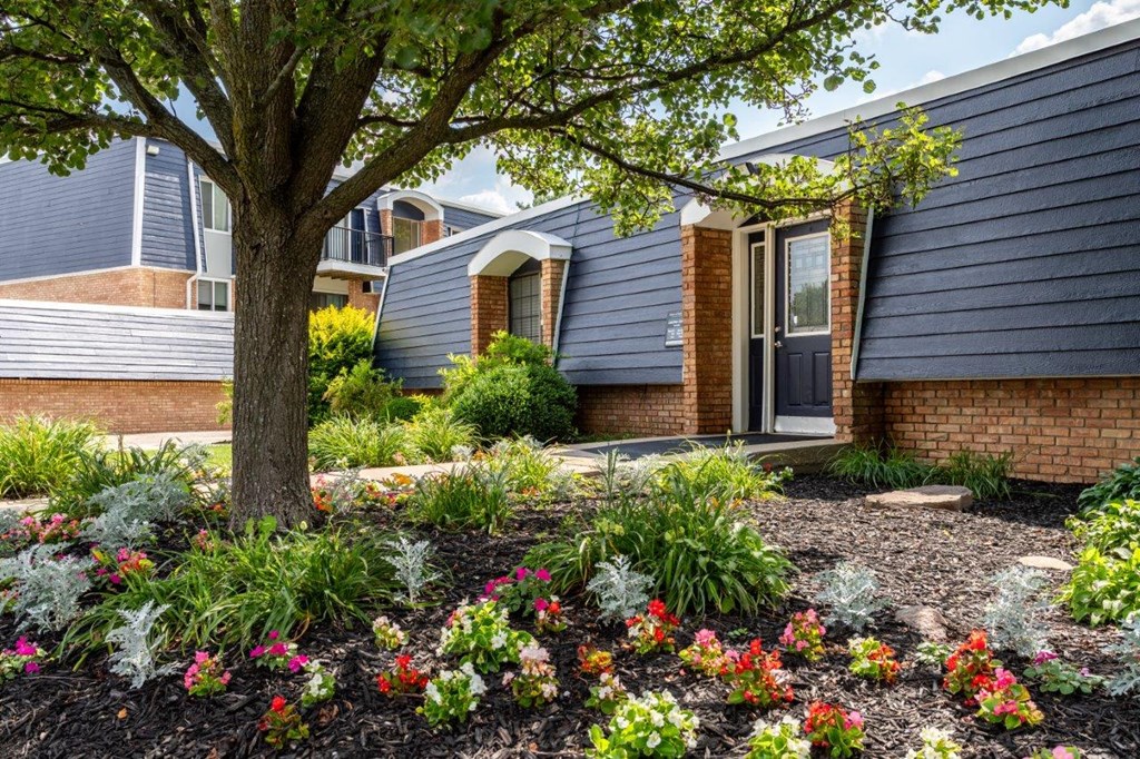 A house with a blue siding and a brick facade has a tree and flowers in front.