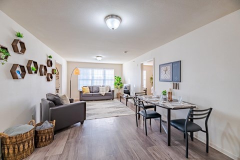 A well-lit living room with a grey sofa, a dining table with chairs, and a wooden floor.