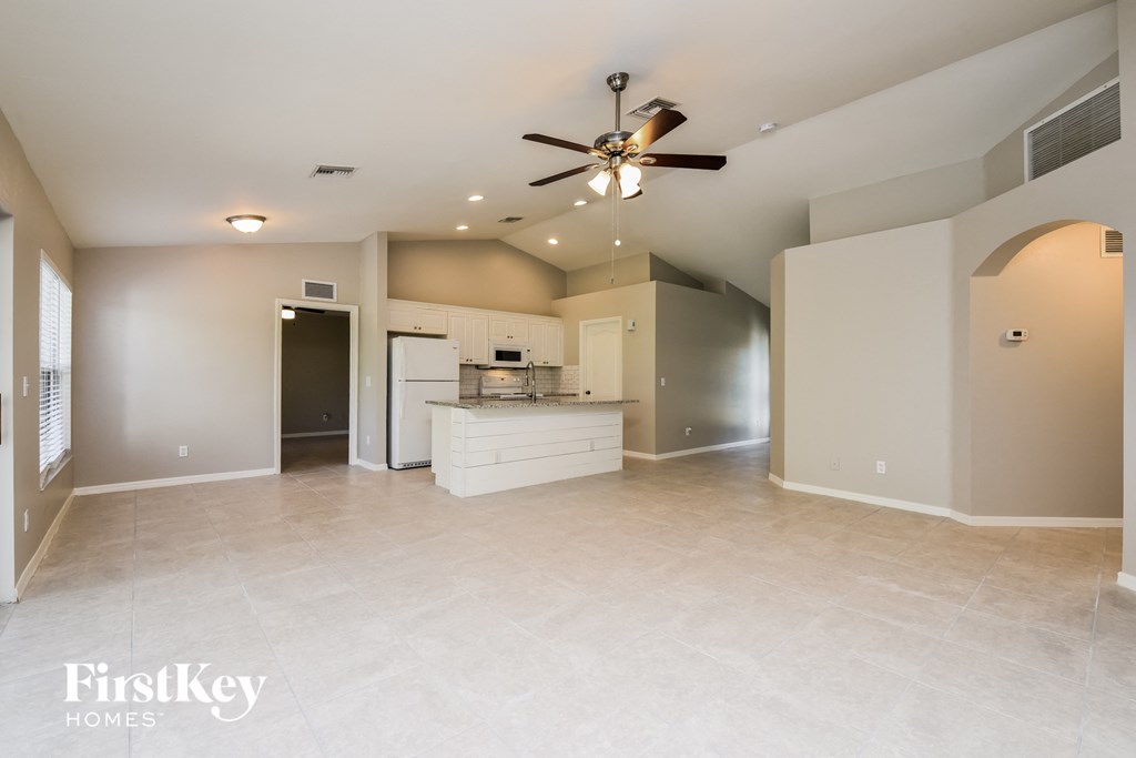 A spacious, empty room with a ceiling fan and a kitchen area in the background.