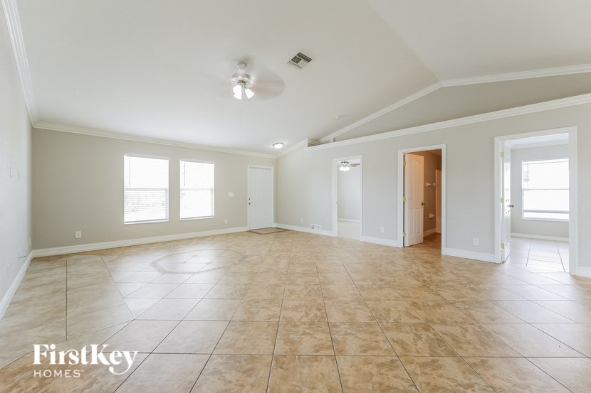 A spacious room with beige tiled flooring and white walls, with a ceiling fan and three windows.