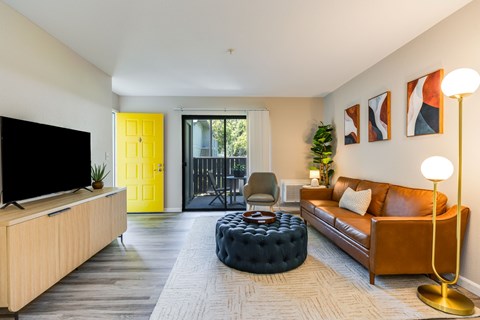 A living room with a brown leather couch, a black ottoman, a wooden cabinet, a flat screen TV, and a yellow door.