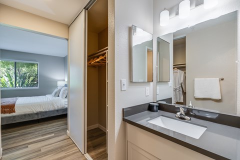A bathroom with a sink, mirror, and a bedroom with a bed and a window.