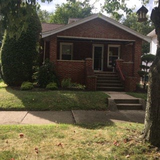 A small red brick house with a front porch and a tree in front.