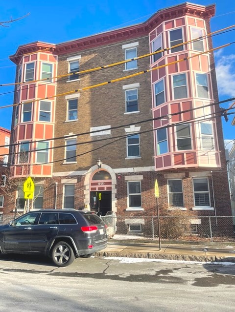 A car is parked in front of a multi-story building with a yellow warning sign on the sidewalk.