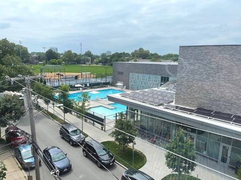 A view of a street with cars parked on the side and a building with a pool in the background.