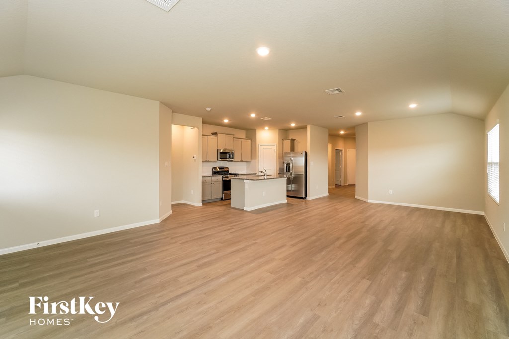 A spacious empty room with wooden flooring and a kitchen area in the background.