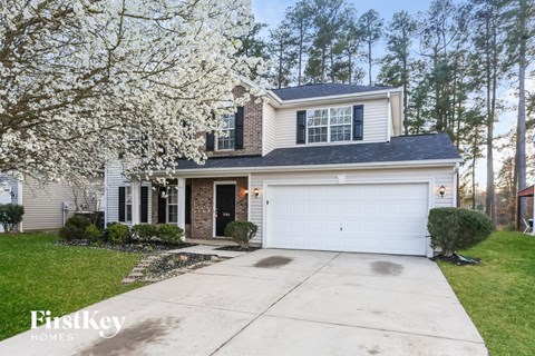 A two-story house with a garage is surrounded by trees and has a white flowering tree in the front yard.