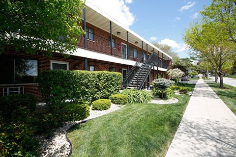 a sidewalk in front of a brick apartment building with grass and bushes
