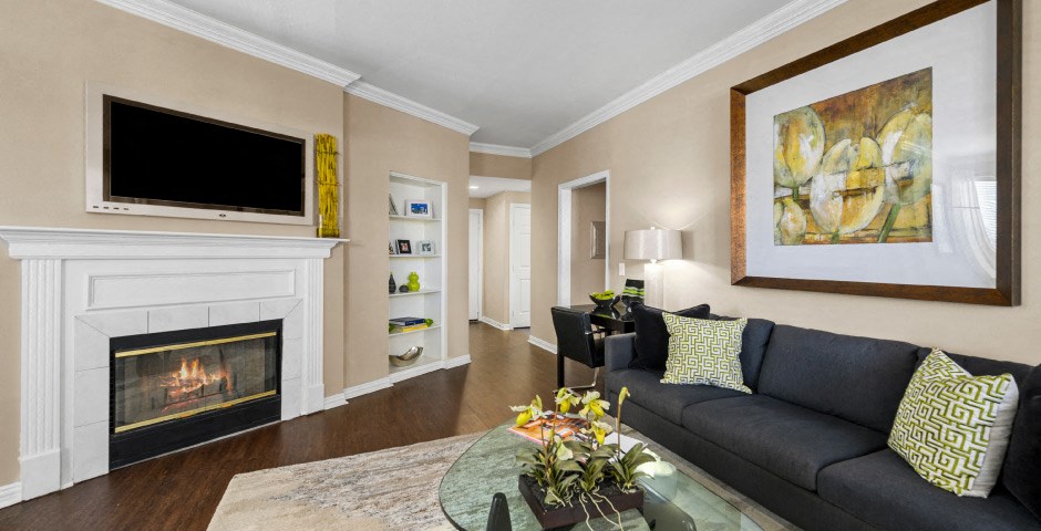 Spacious living room with wood-burning fireplace with glass doors, ceiling fan, 9-foot ceilings, built-in shelves and wood grain plank flooring at The Lanesborough Apartments in Houston.