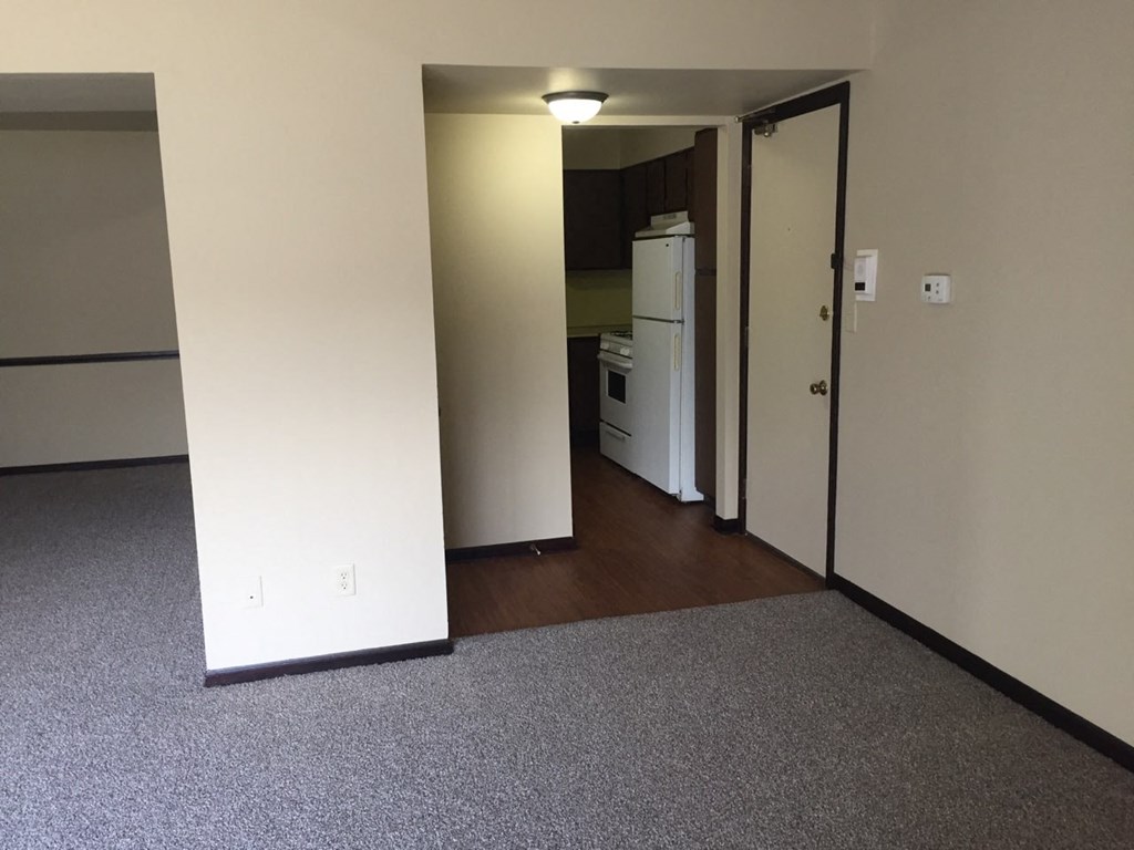 an empty living room with a refrigerator and a door to a kitchen