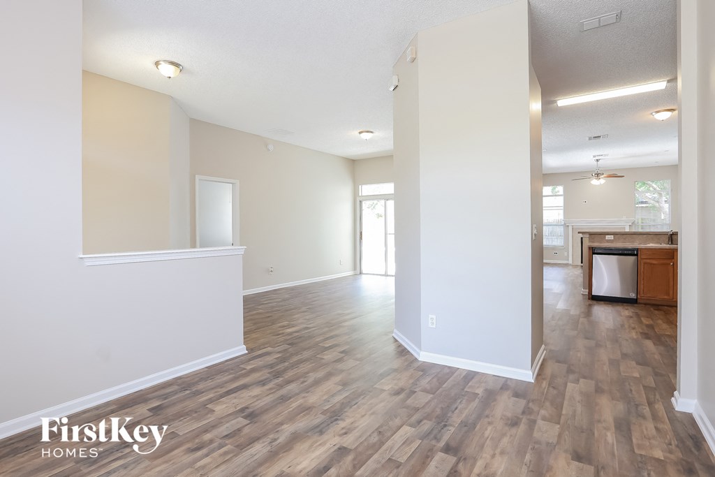 A spacious room with wooden flooring and white walls, with a kitchen area visible in the background.