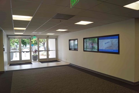 A hallway with a green exit sign above the door and a monitor on the wall.