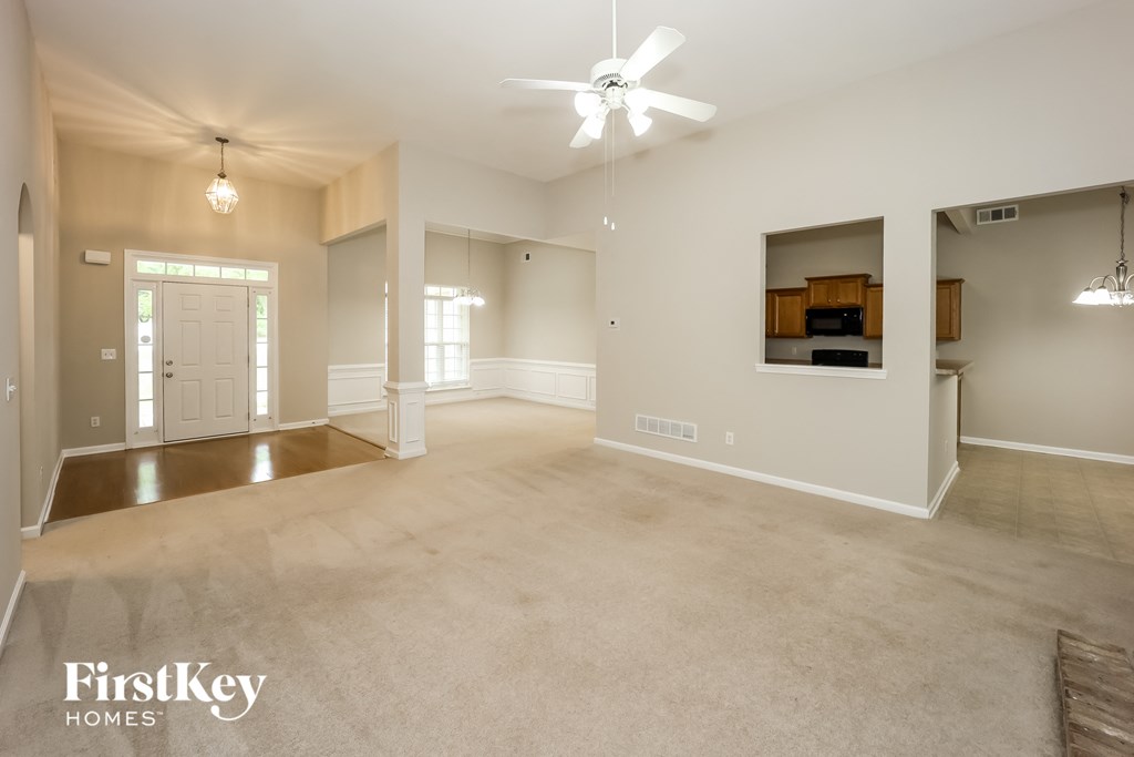 A spacious living room with a ceiling fan and a view of a kitchen in the background.