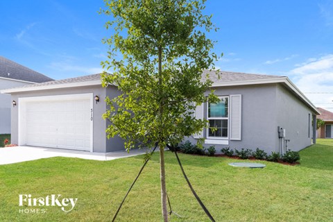 A tree stands in the front yard of a grey house with a white garage door.
