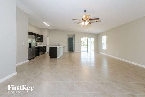 A spacious empty room with a ceiling fan and a kitchen area in the background.