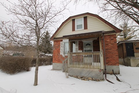 A small house with a porch and a fence is surrounded by snow.