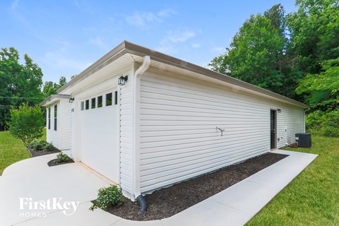 A white house with a brown roof and a garage door is surrounded by greenery.