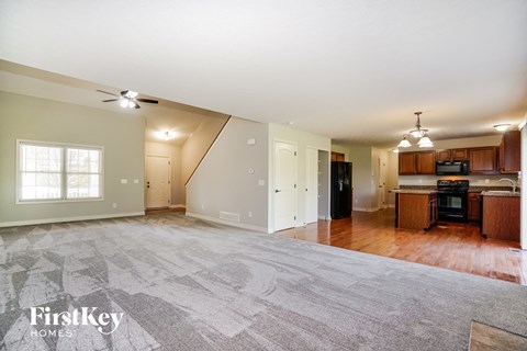 A spacious living room with a grey carpet and a kitchen area with wooden cabinets.