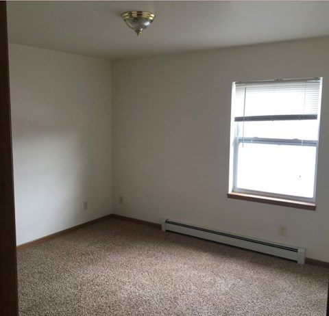 A room with a carpeted floor, a window with blinds, and a ceiling light fixture.