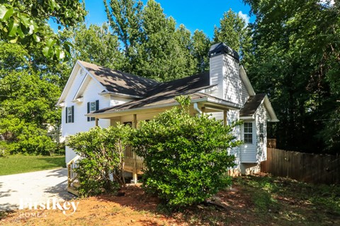 A white house with a black roof and a chimney is surrounded by greenery.