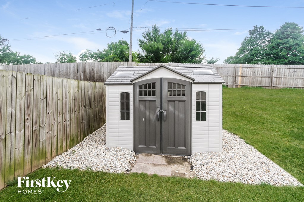 A small white shed with a grey door is situated in a grassy area with a wooden fence in the background.