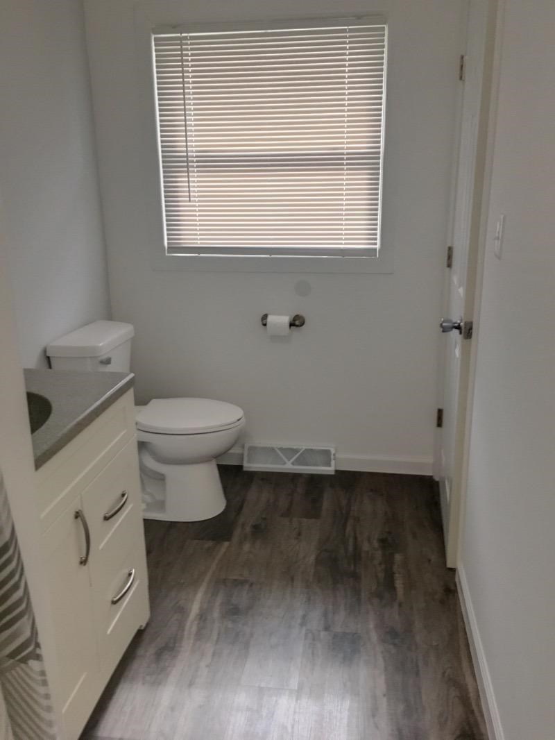 A white toilet sits in a bathroom with a wooden floor and a window with blinds.