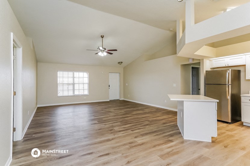 an empty living room with a kitchen and a stainless steel refrigerator