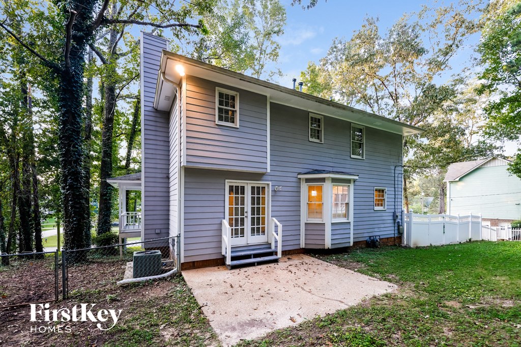 A small house with a porch is surrounded by trees and has a fence in the front yard.