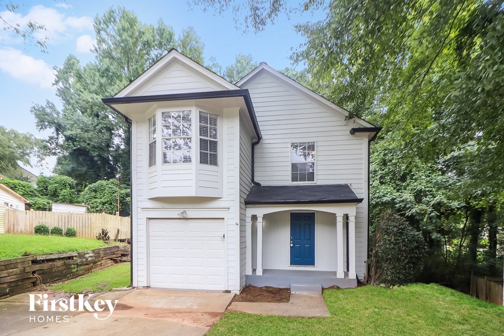 A white house with a black roof and a blue door is surrounded by greenery.