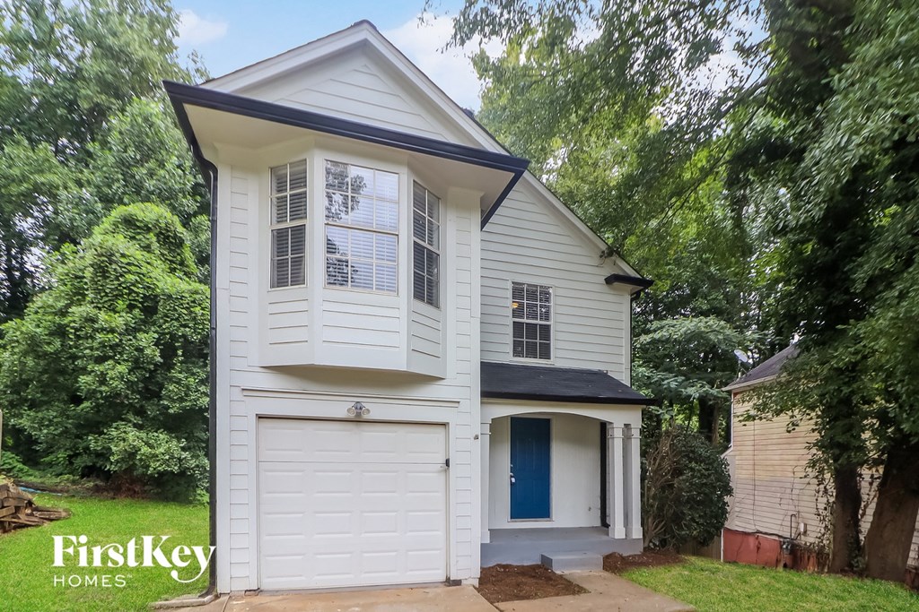 A white house with a black roof and a blue door is surrounded by greenery.