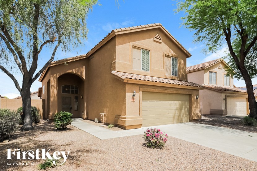A house with a brown stucco exterior and a tan roof is surrounded by trees and has a gravel driveway.