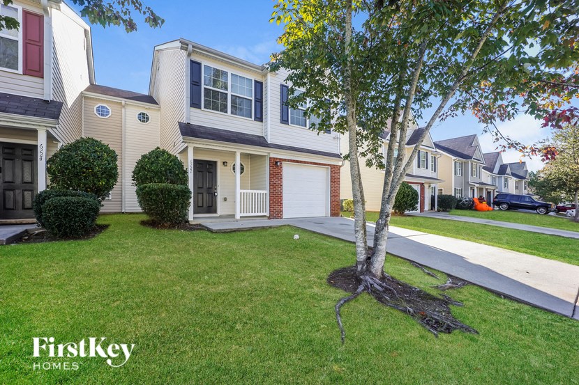 A tree with a thick trunk and a small branch is planted in a grassy area in front of a house.