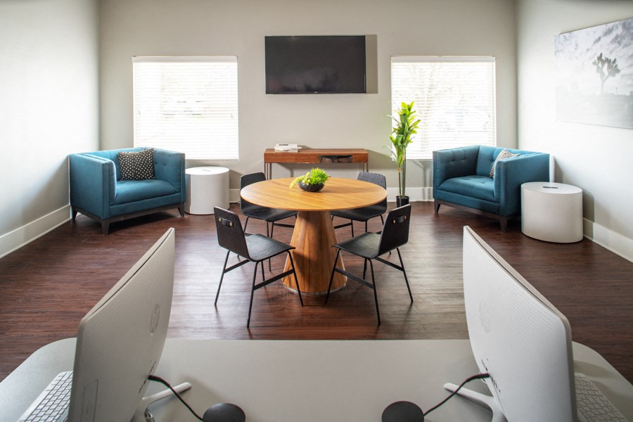A modern office with a wooden table surrounded by blue chairs and two computer monitors.