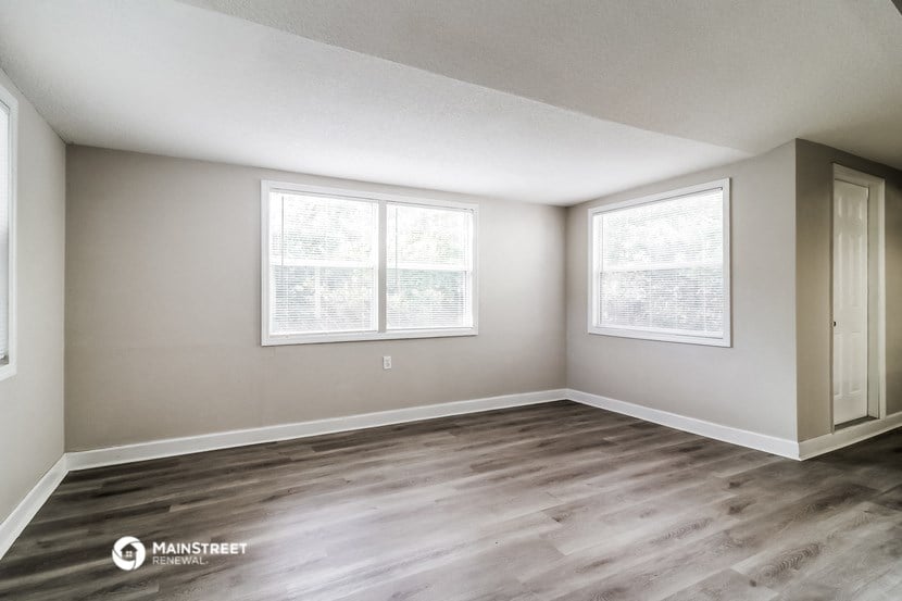 the spacious living room of an apartment with wood flooring and two windows