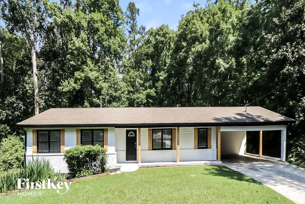 A white house with a brown roof and a black door is surrounded by greenery.