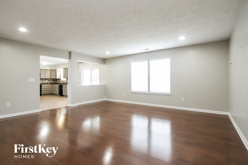 A spacious room with wooden floors and white walls, with a view of a dining area in the background.