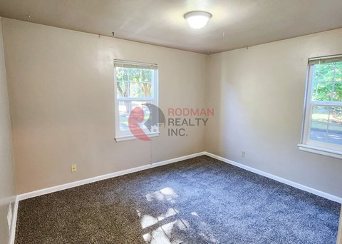 A room with a carpeted floor and a window with a view of trees and a building outside.