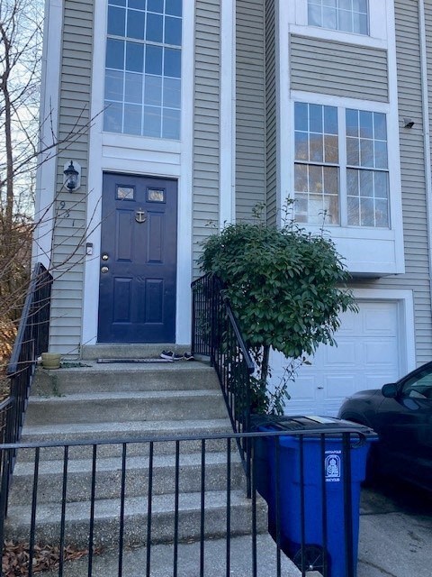 A blue trash can is in front of a house with a black car parked in front of the garage.
