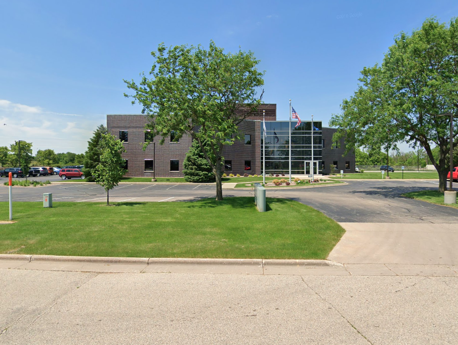 A modern building with a flag on top is surrounded by trees and a parking lot.