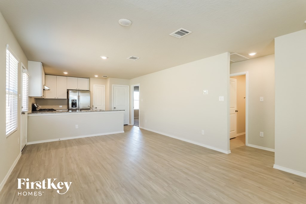 A spacious, empty room with wooden floors and white walls, with a kitchen area visible in the background.