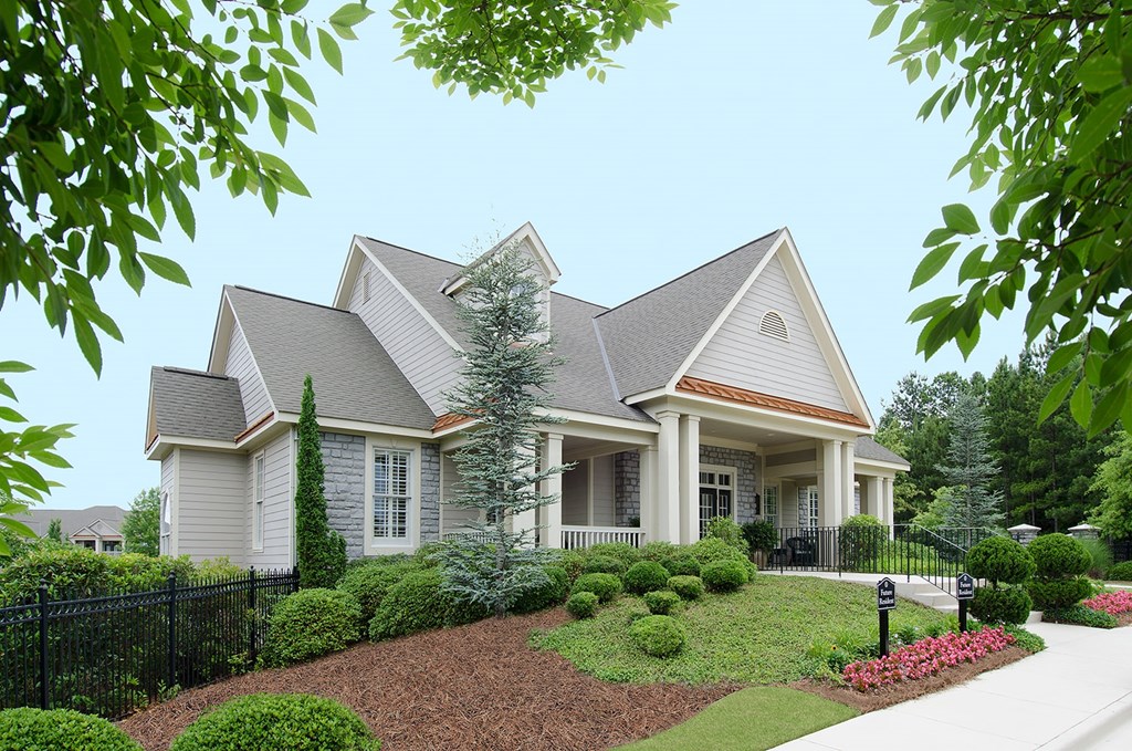A house with a grey roof and white walls is surrounded by a black fence and green bushes.