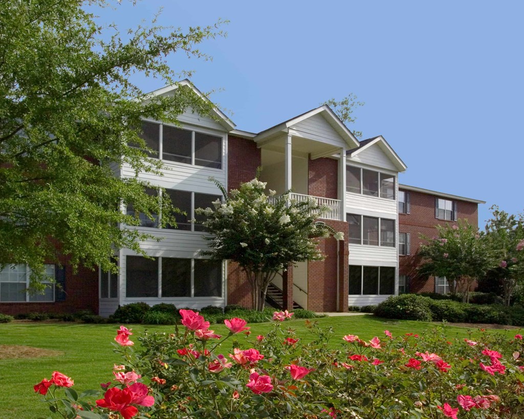 A building with a red brick facade and white trim is surrounded by a green lawn and red flowers.