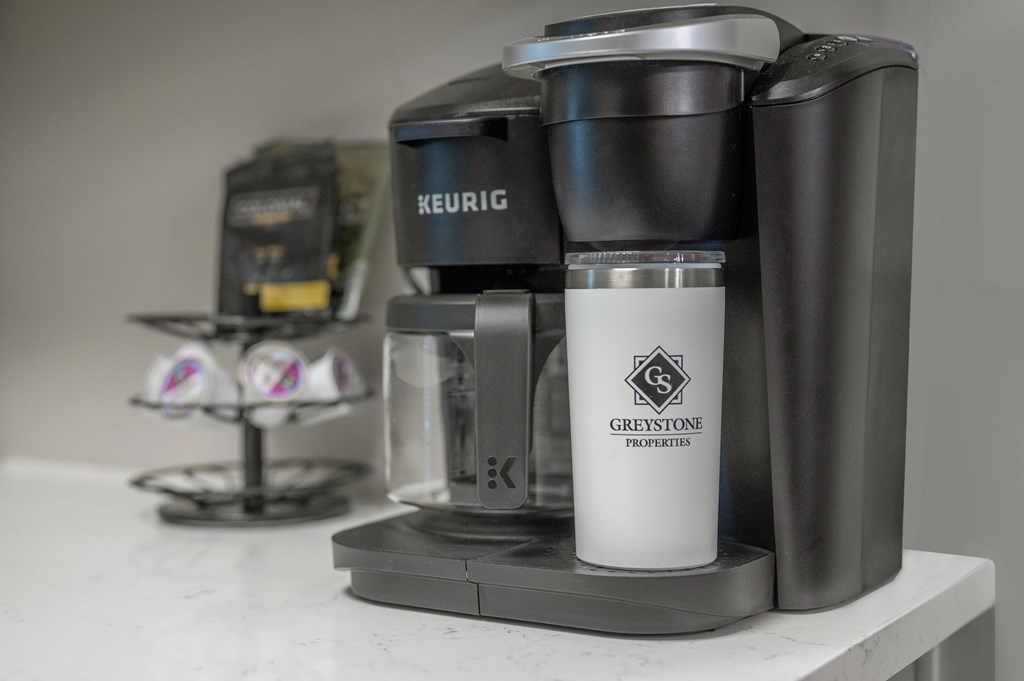 A Keurig coffee maker is on a counter with a Greystone Properties mug in front of it.
